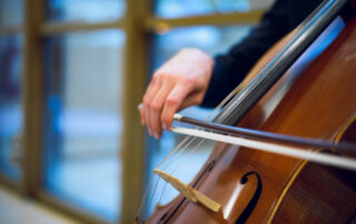close up of a hand drawing a cello bow across the cello strings in front of a row of windows