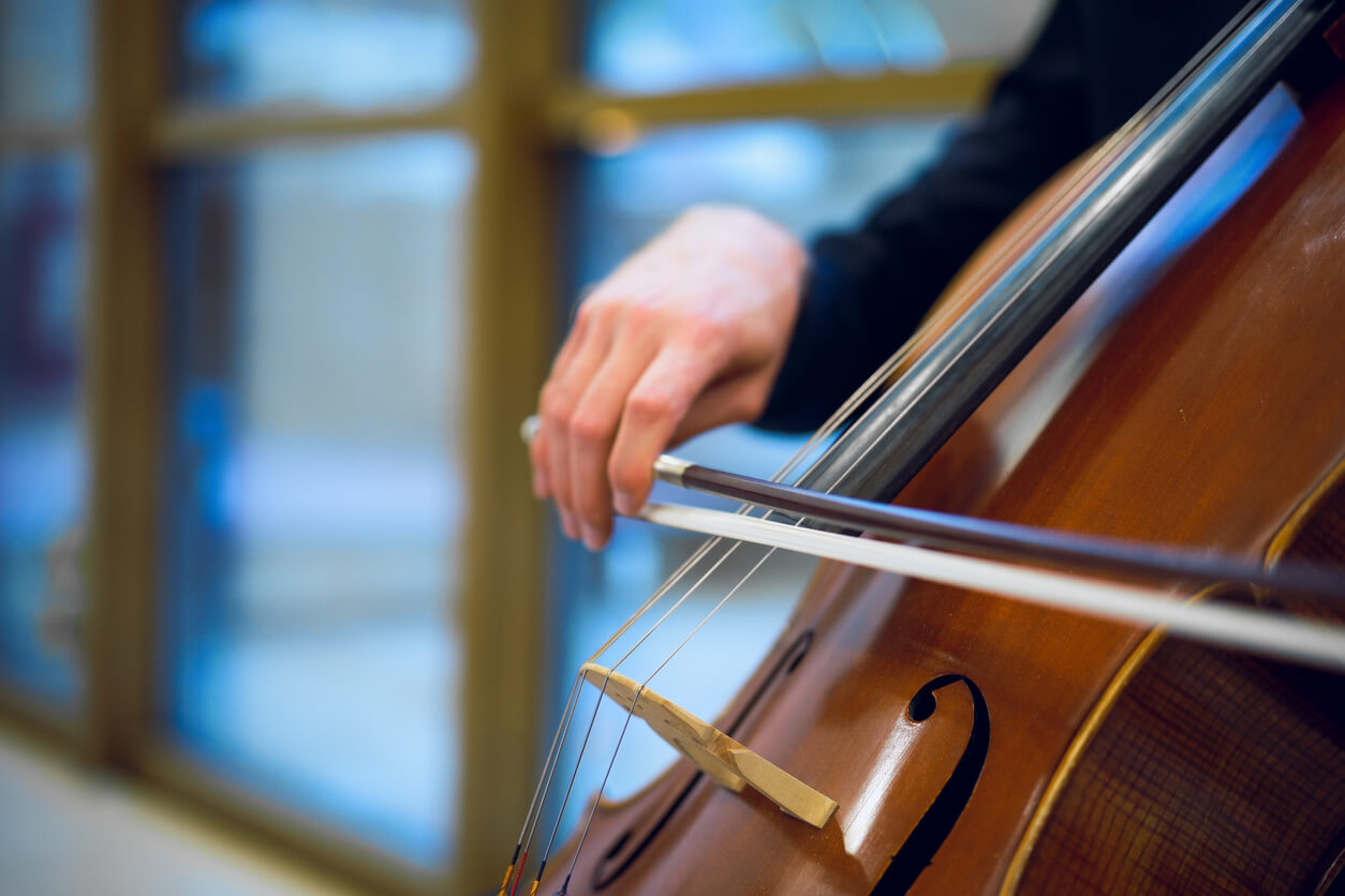 close up of a hand drawing a cello bow across the cello strings in front of a row of windows