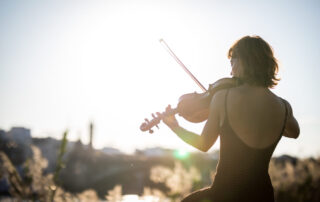 back of a woman in a black dress playing violin outside with the sun shining brightly in front of her