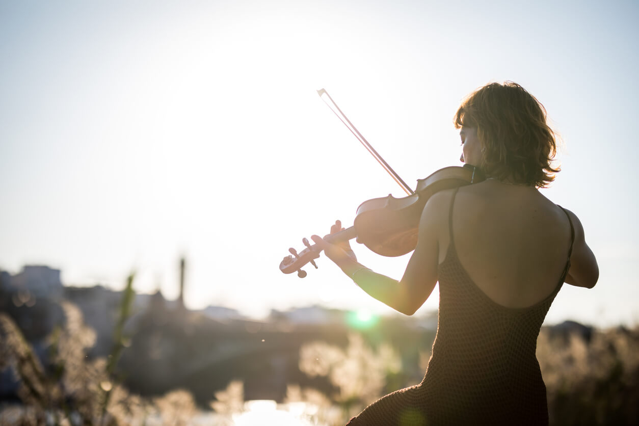 back of a woman in a black dress playing violin outside with the sun shining brightly in front of her