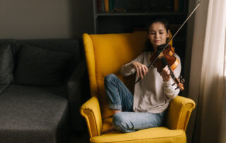 young woman casually sitting in a yellow living room chair and playing the violin