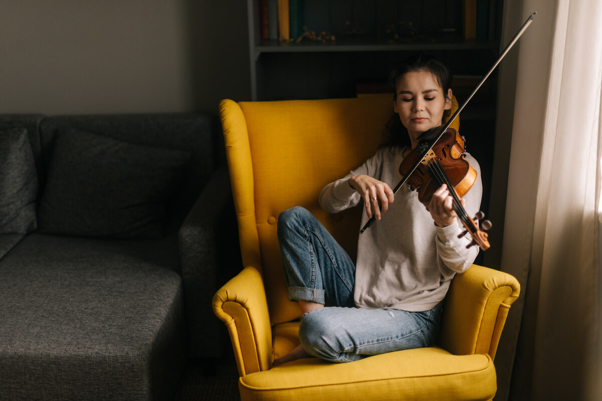 young woman casually sitting in a yellow living room chair and playing the violin