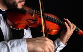 man with facial hair in a dress shirt playing a violin against a black background