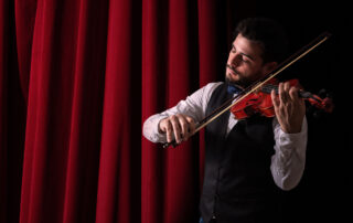 Man in button up and vest playing a violin in front of a red curtain