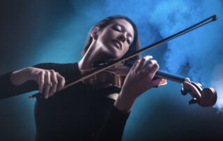 woman in black clothing passionately playing the violin in front of a smoky, black background