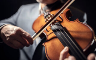older violinists hands holding and playing violin with violin in focus and man's body in background out of focus