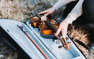 woman's hands placing a violin bow in a case that already has a bow inside. the case is on a ground covered in dry grass.