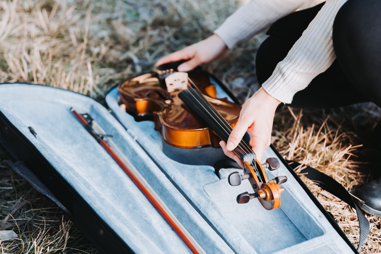 woman's hands placing a violin bow in a case that already has a bow inside. the case is on a ground covered in dry grass.